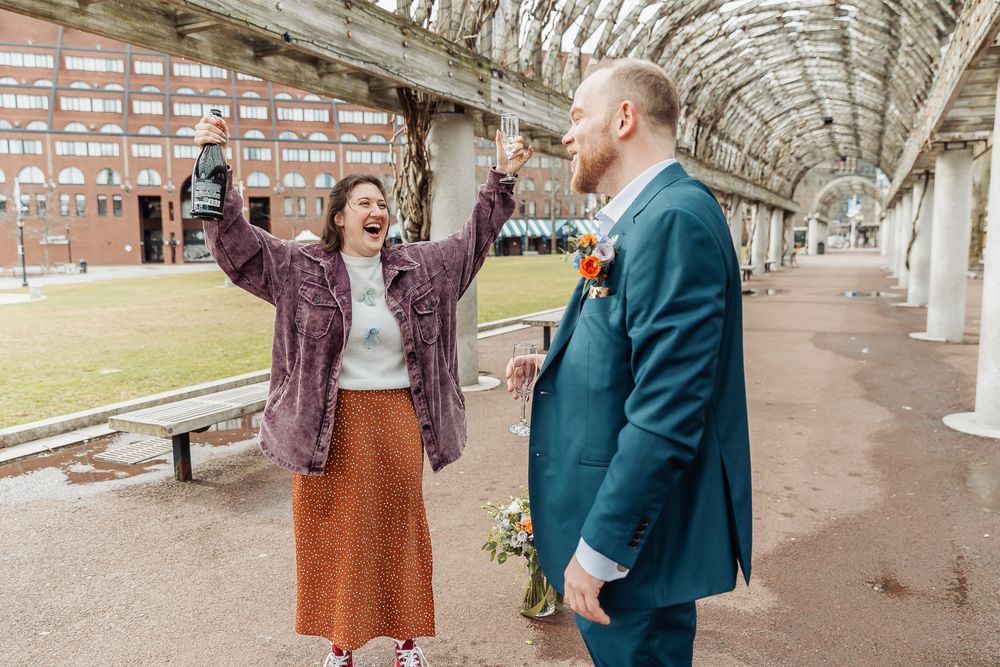 Joyful bride celebrating with raised arms at outdoor venue while groom holds bouquet.