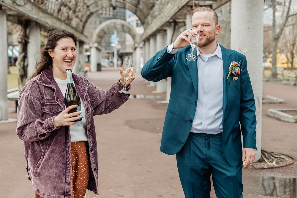 Joyful couple celebrating outdoors, woman in denim jacket holding beer bottle, man in teal suit drinking, under stone arches.