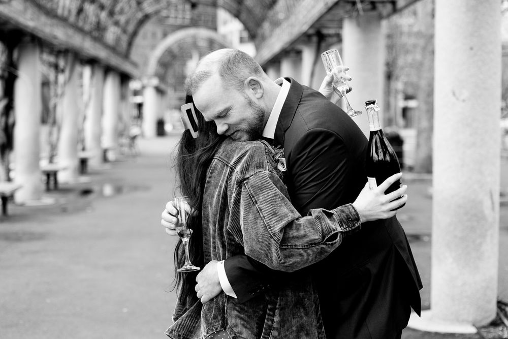 Two men share an emotional embrace in black and white photo, holding glasses at a formal outdoor event.