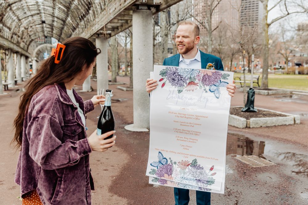 Couple celebrating outdoors under pergola, man holding large floral wedding menu sign, woman with wine bottle.