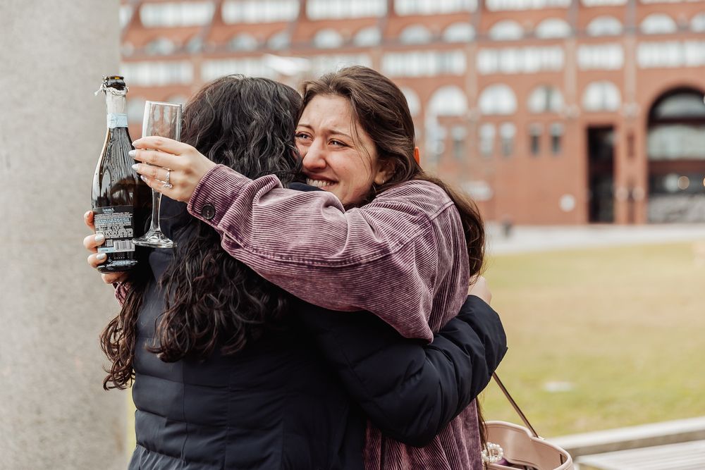 Two women sharing an emotional hug while holding champagne bottle and glass outdoors.