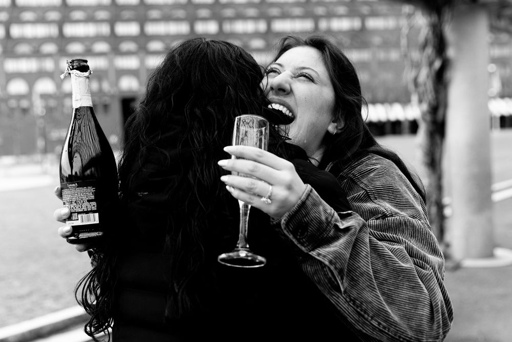 Two women celebrating outdoors, one biting the other playfully while holding champagne glasses in black and white.