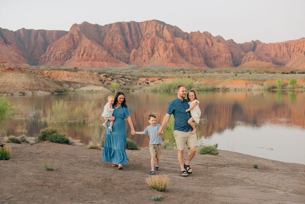 Family of five walking hand-in-hand along a scenic desert lake with red rock cliffs reflecting in calm water.