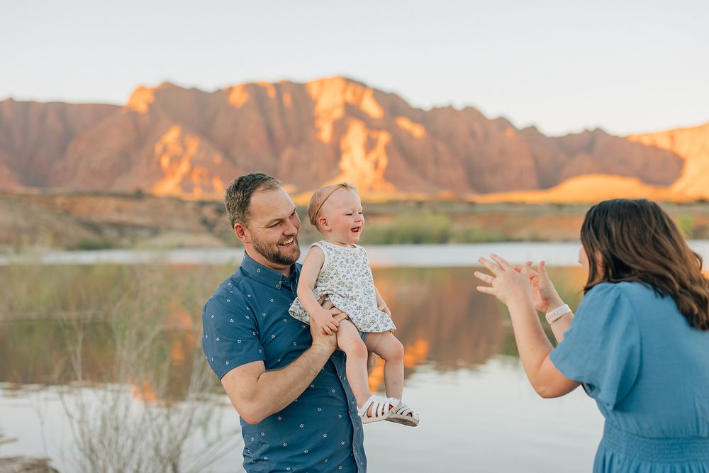 Family enjoying golden hour by a lake with red rock mountains, father holding laughing baby while mother plays.