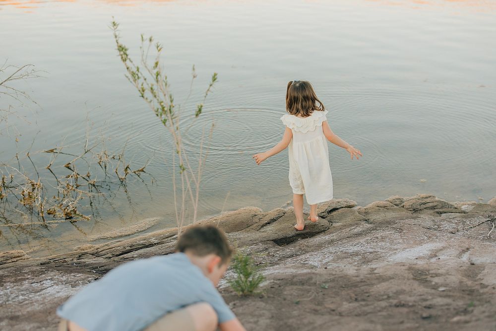 A young girl in a white dress walks along rocks near calm water, holding a rope while a person crouches nearby.