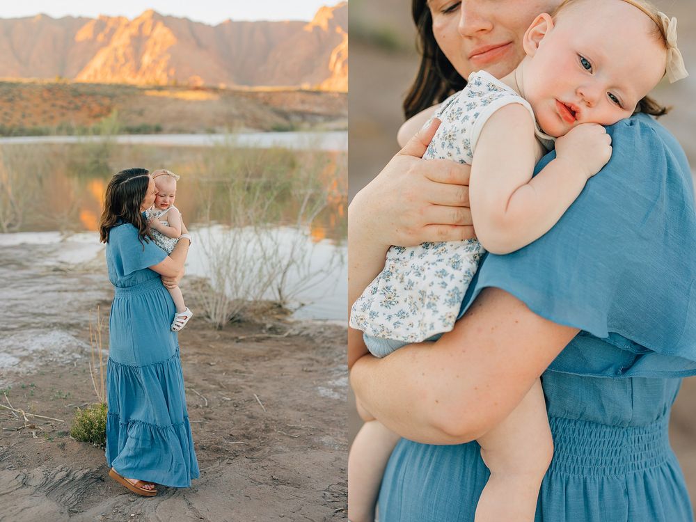 Mother in blue dress holds smiling baby outdoors near sandy riverbank with golden mountain backdrop at sunset.