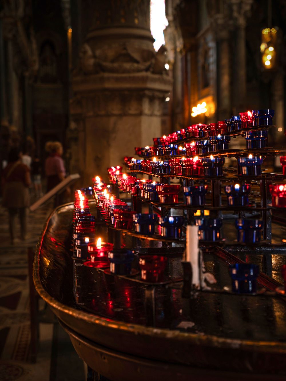 Red and blue votive candles glowing in a ornate holder inside a dimly lit cathedral.