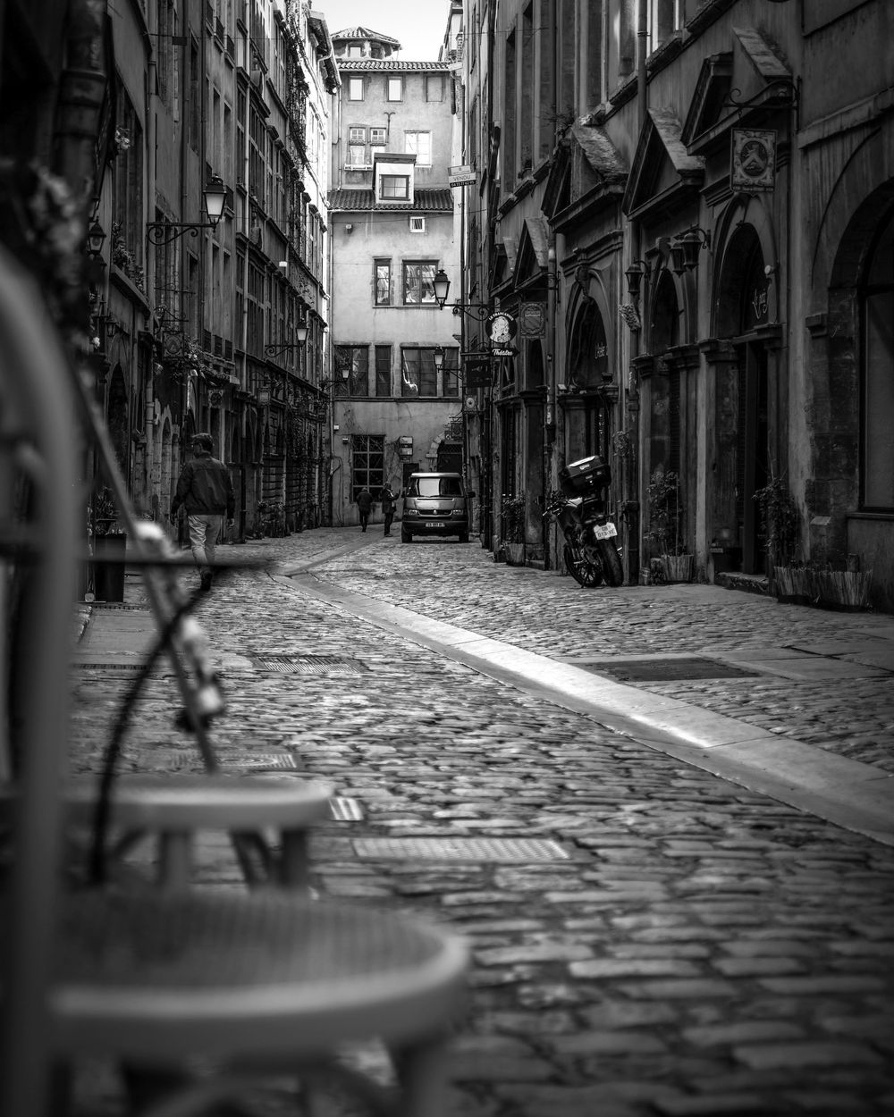Black and white photo of a cobblestone alley in an old European city with buildings and a parked motorcycle.