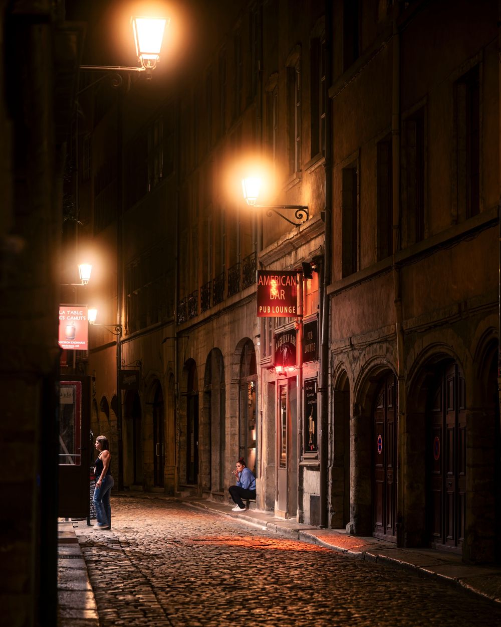 A cobblestone alley at night in Lyon, France, lit by warm glowing street lamps with a lone figure standing nearby.