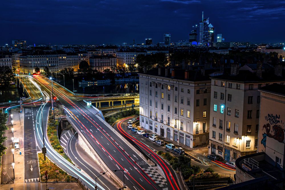 Aerial night view of a busy city highway with light trails from traffic, surrounded by urban buildings and skyline.
