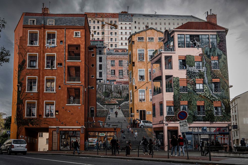 Colorful trompe-l'oeil mural on building facades in Lyon, France, depicting realistic architecture with pedestrians below.