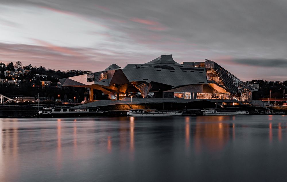 Modern architectural building illuminated at dusk reflecting on calm water with dramatic cloudy sky.