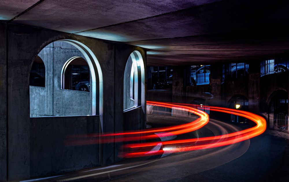 Long exposure photo of red and orange car light trails curving under a dark concrete underpass at night.