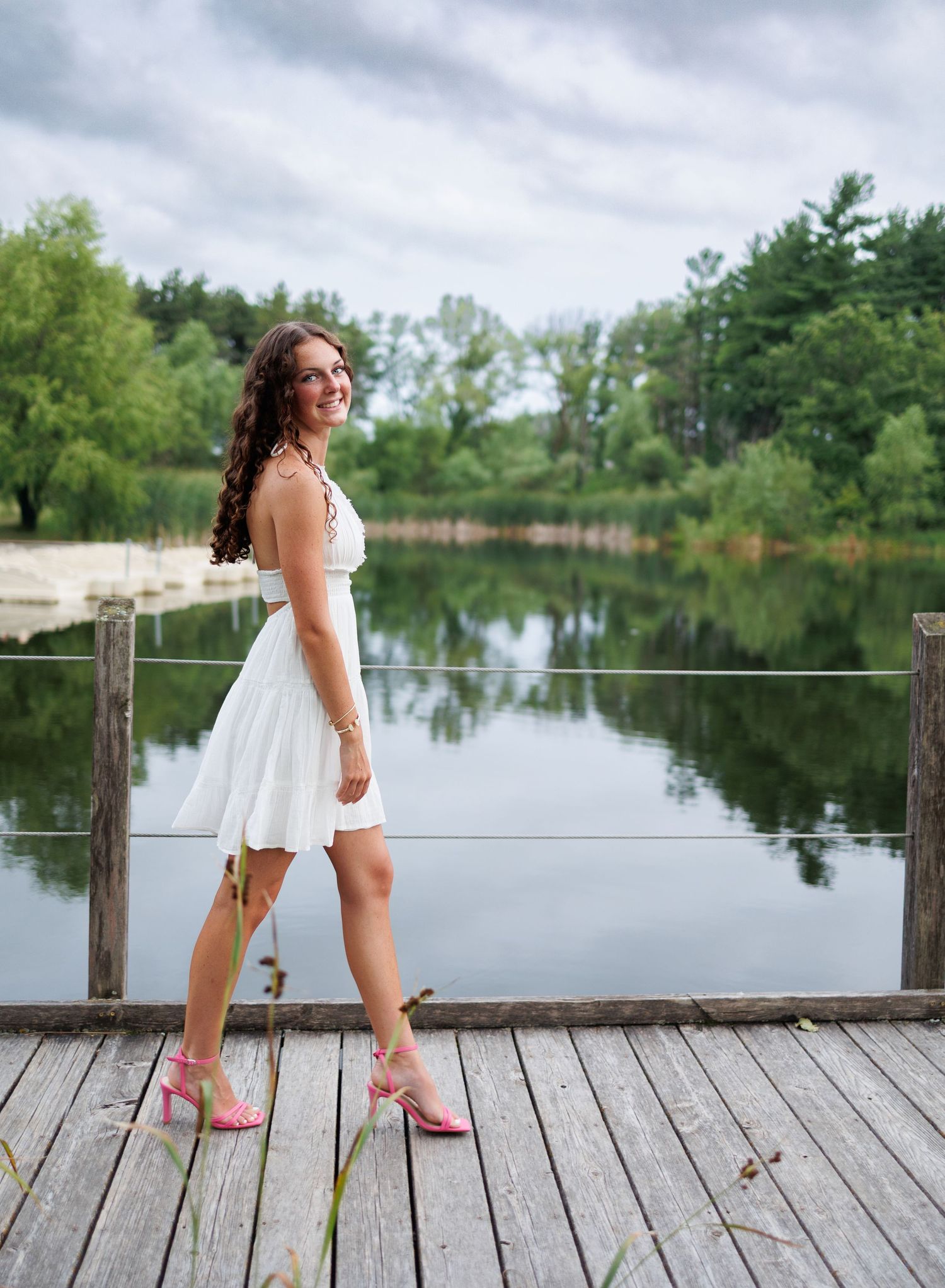 A senior girl in a white summer dress poses playfully on a wooden dock overlooking a serene lake on a cloudy day.