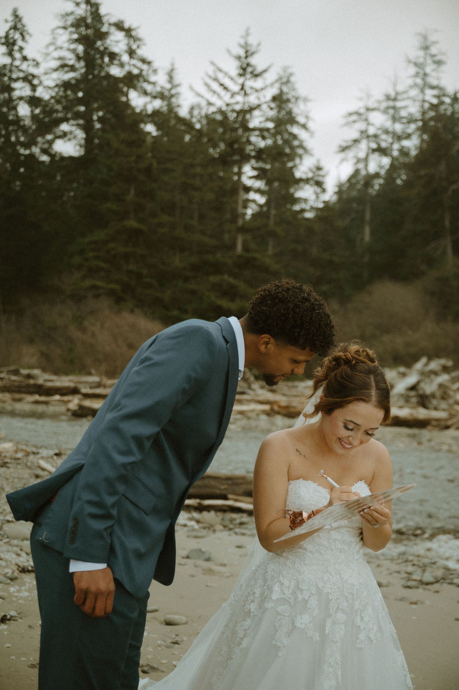 A couple shares a tender moment on a rocky beach in formal wedding attire against a backdrop of evergreen trees.