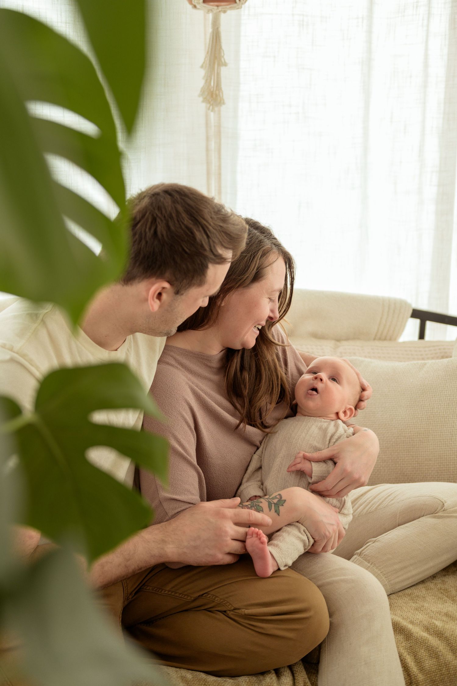 A couple shares a tender moment while relaxing together on a beige couch near a window with green plants.