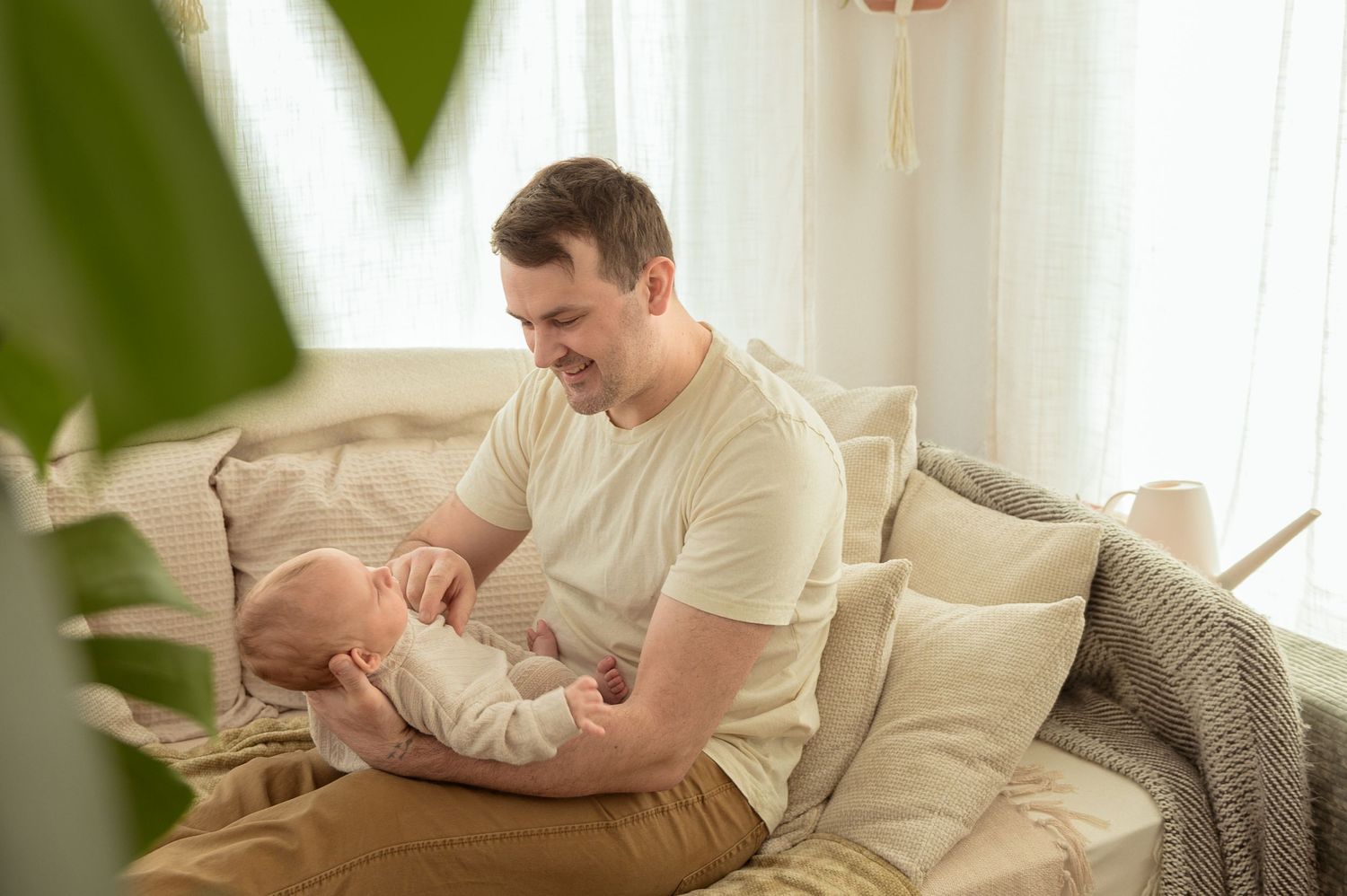 A parent sits on a beige couch holding a small baby while framed by houseplants and natural window light.