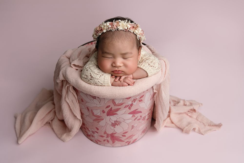 Newborn baby sleeping peacefully in pink floral bowl with soft beige wrap and delicate flower headpiece.