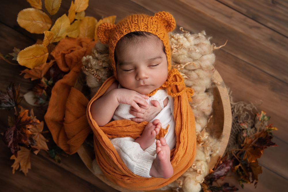 Newborn sleeping peacefully in orange wrap and bonnet surrounded by autumn leaves and floral decorations on wooden floor.