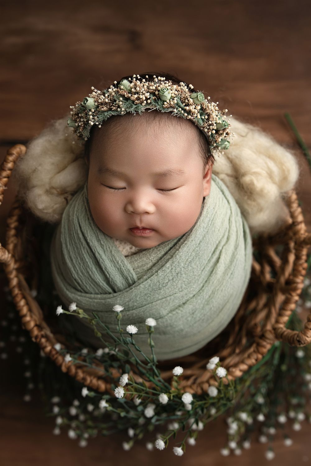 A sleeping newborn wrapped in soft sage green fabric rests in a rustic basket adorned with delicate baby's breath flowers.