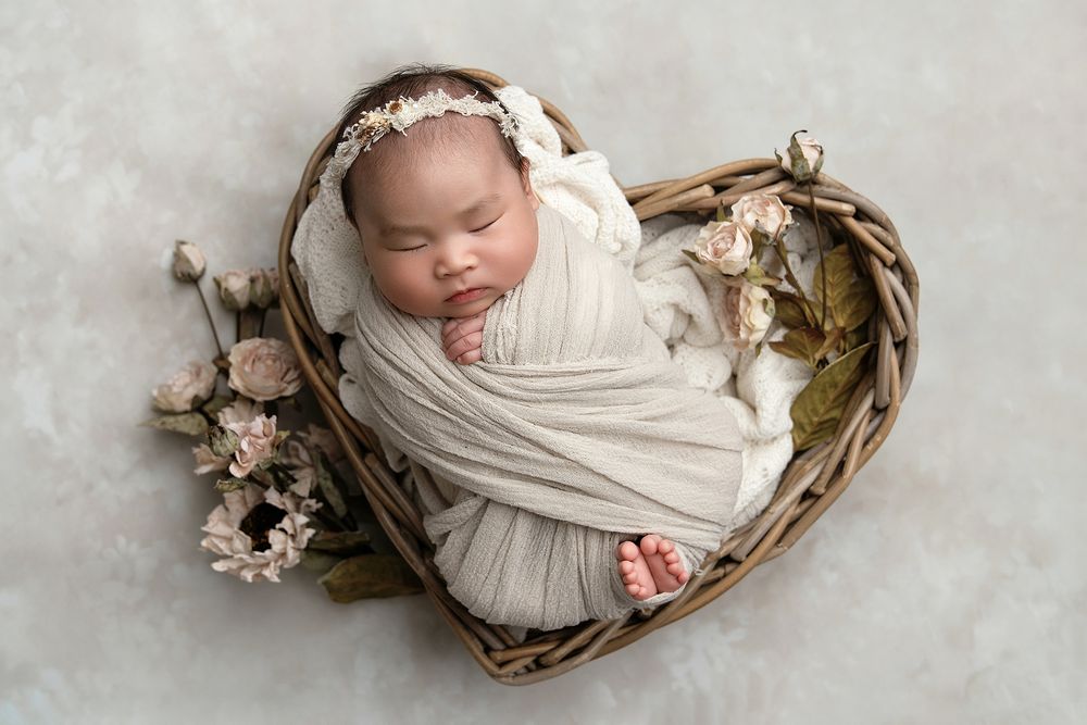 A sleeping newborn baby swaddled in white fabric lies peacefully in a rustic wooden basket with delicate floral accents.