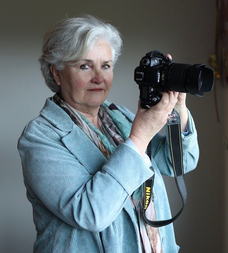 Elderly woman with gray hair holding a Nikon camera, wearing a teal blazer and colorful scarf.