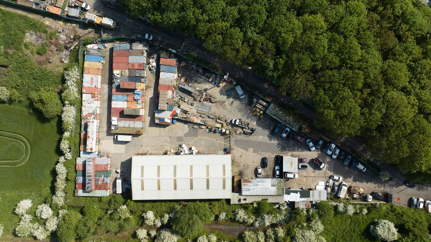 Aerial view of an industrial yard with shipping containers, machinery, and warehouses surrounded by lush green trees.