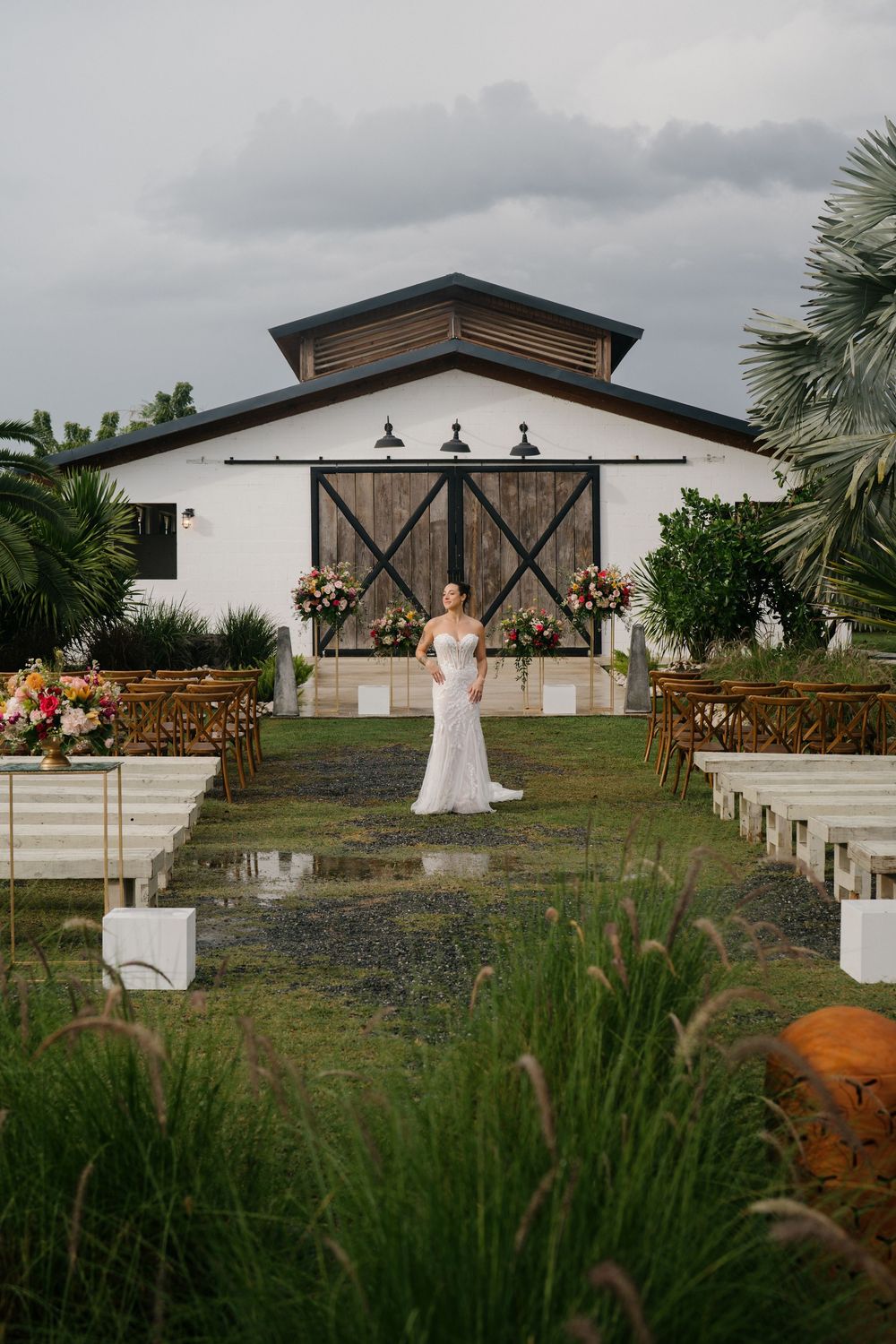 Modern barn wedding venue with rustic black doors and white exterior surrounded by natural landscaping and wildflowers.