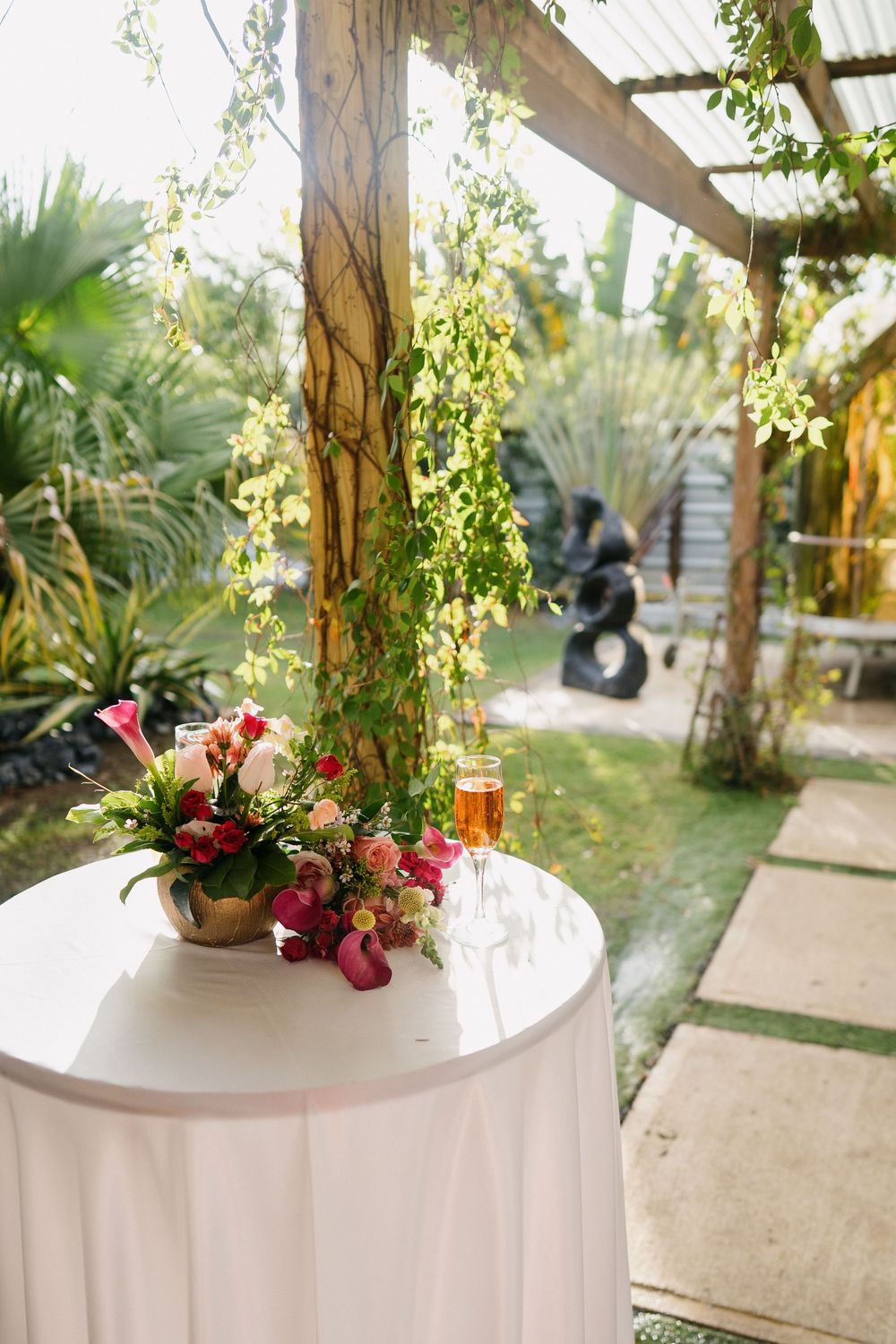 Floral centerpiece on white tablecloth with tropical garden setting and pergola in background.