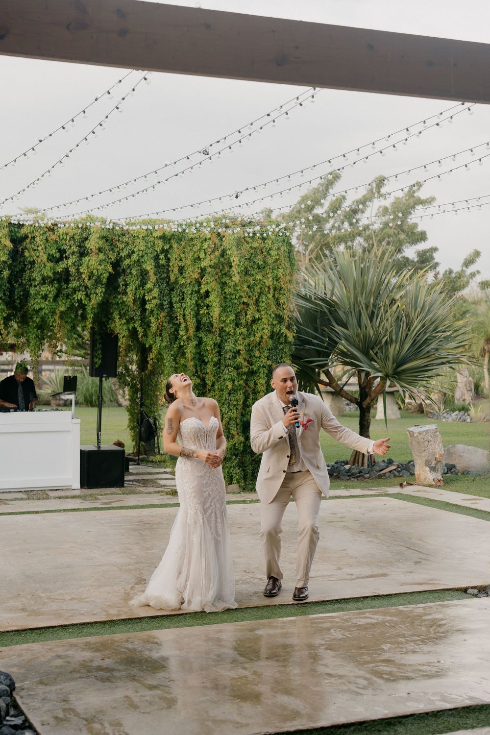 A couple shares a joyful moment dancing together on a wet patio with string lights and greenery in the background.