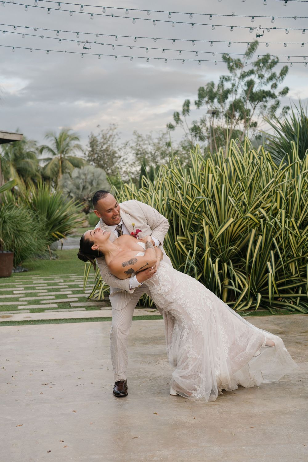 A couple dances together on a patio with tropical plants and string lights overhead during an outdoor celebration.