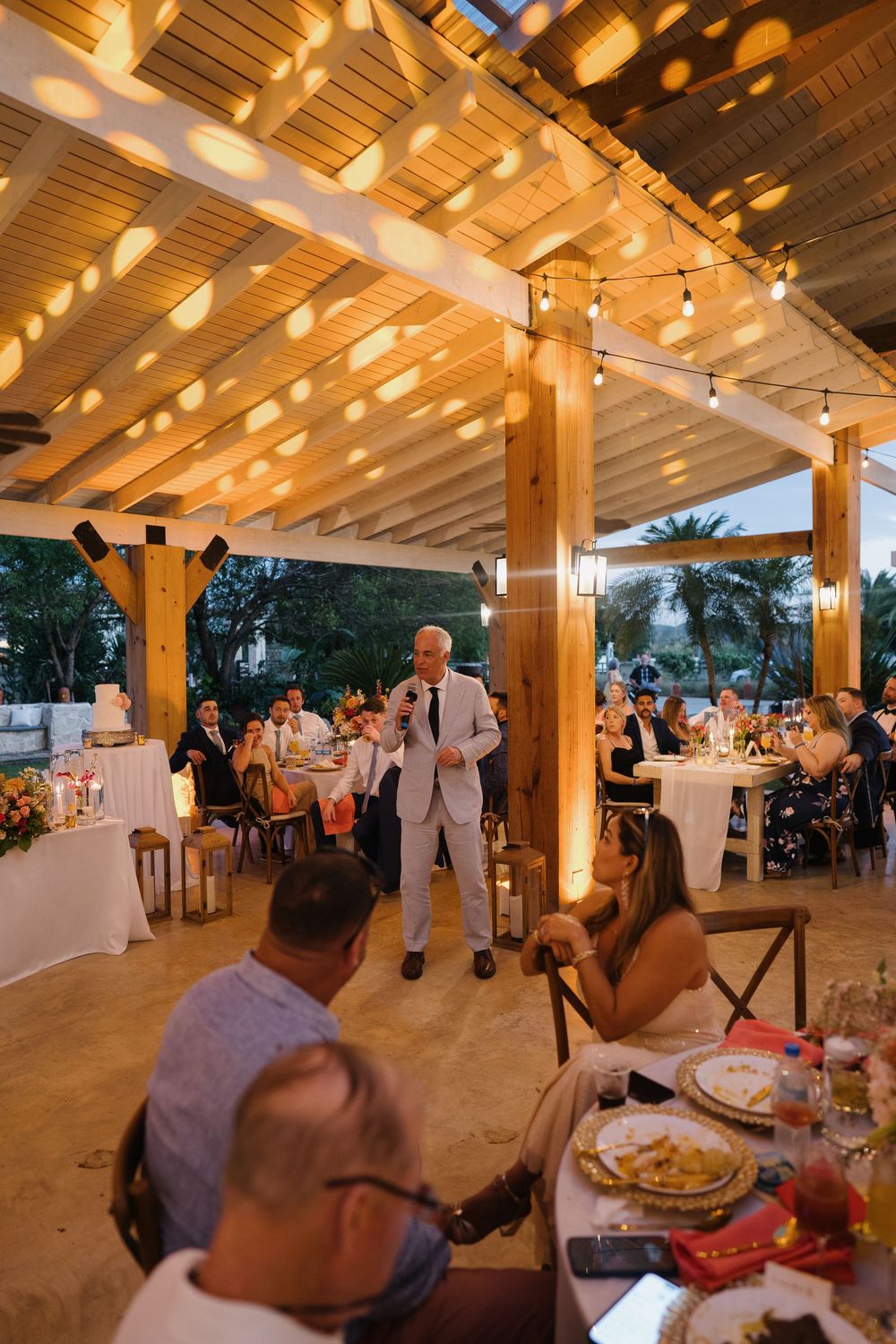 Outdoor wedding reception under a wooden pergola with string lights creating a warm, romantic atmosphere at dusk.
