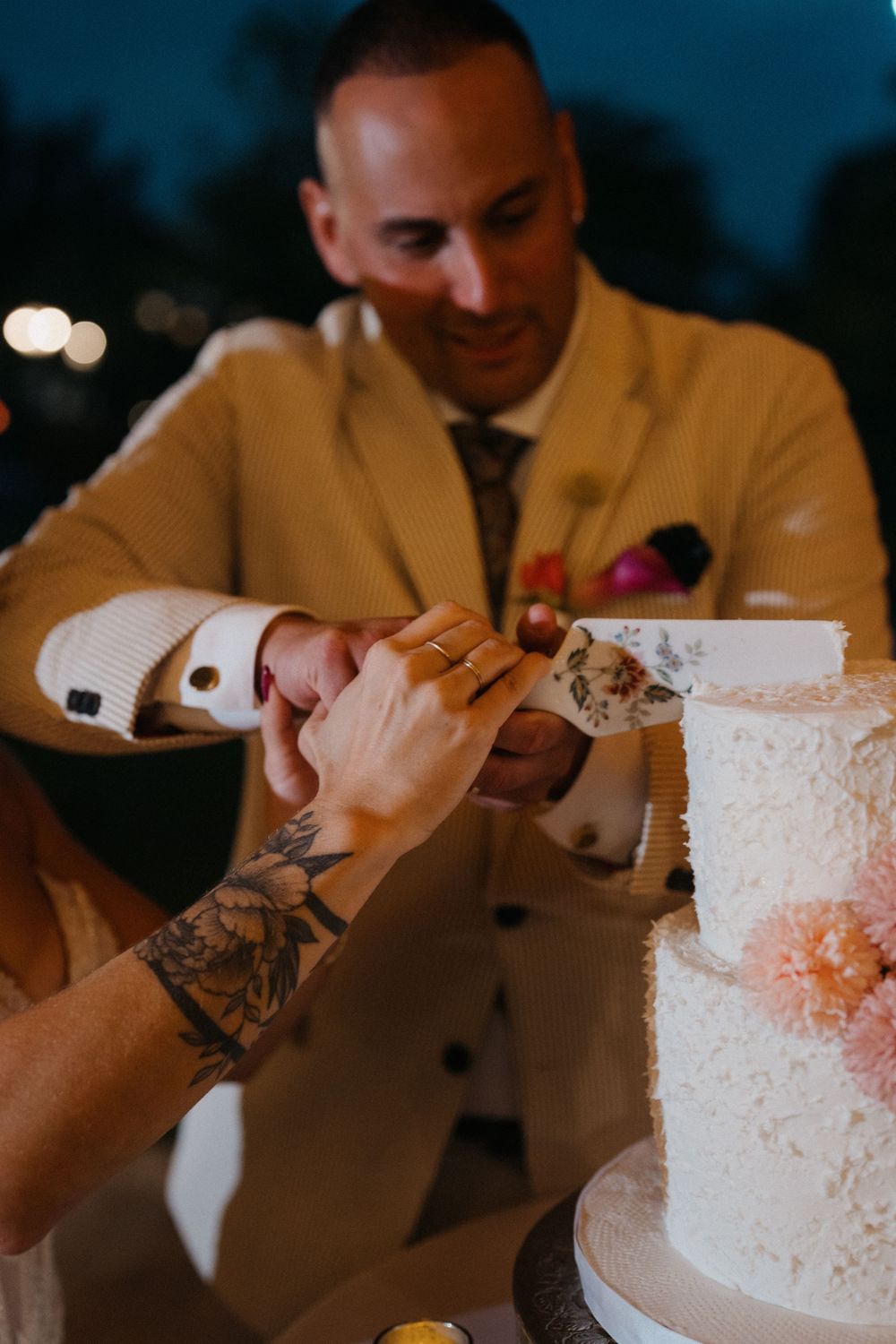 A couple cuts their elegant white wedding cake together at an evening reception, with one person wearing a tan suit.