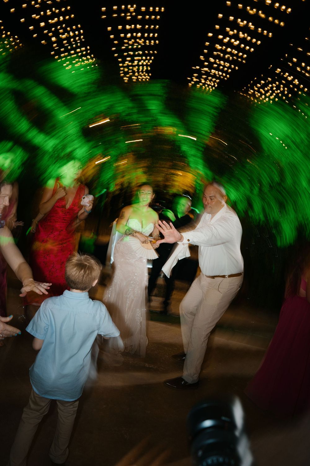 People dancing at a wedding reception under twinkling string lights with palm fronds casting green shadows.