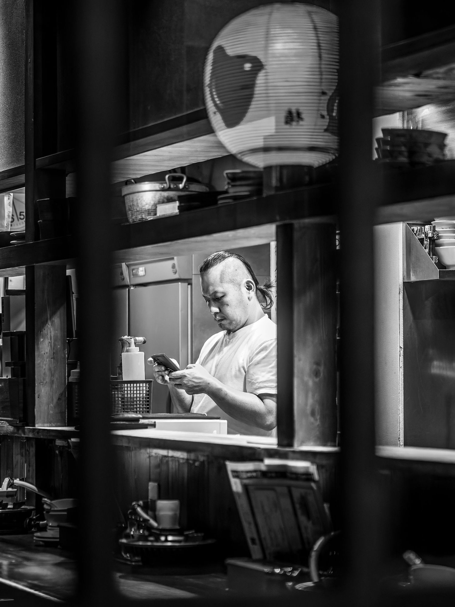 Black and white photo of a chef checking his phone in a traditional Japanese restaurant kitchen.