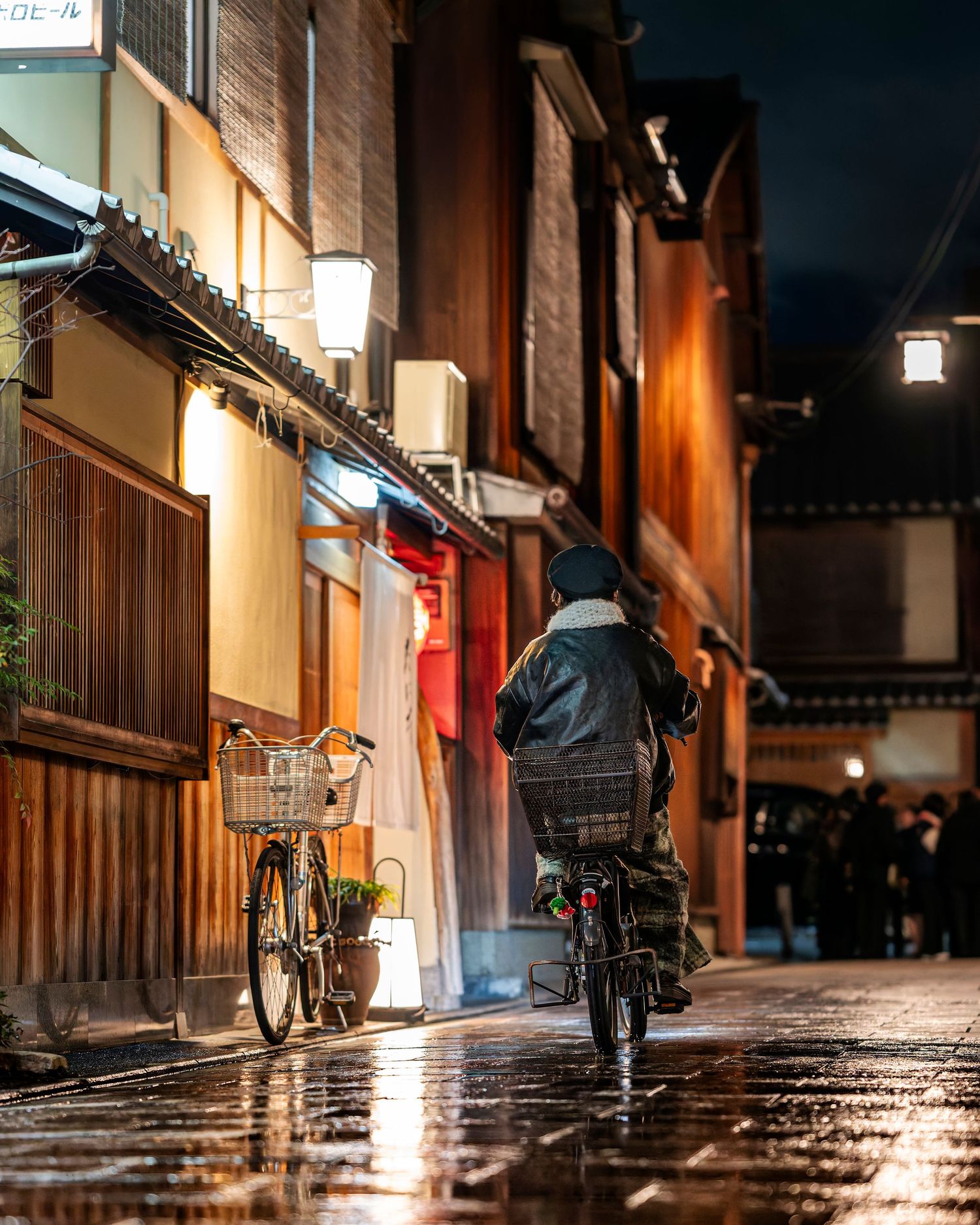 Person cycling through a rain-soaked Japanese alley at night, with warm glowing lanterns and traditional wooden buildings.