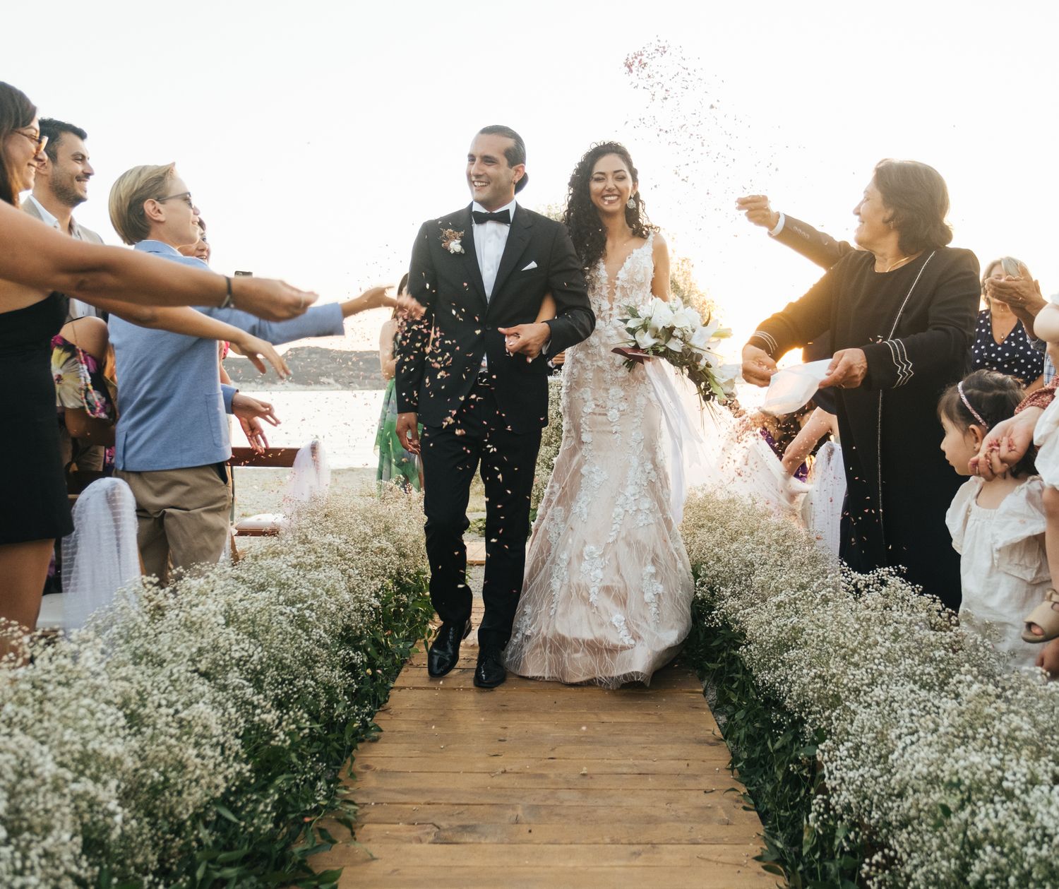 Bride and groom walk down floral aisle as guests toss confetti at outdoor waterfront wedding ceremony.
