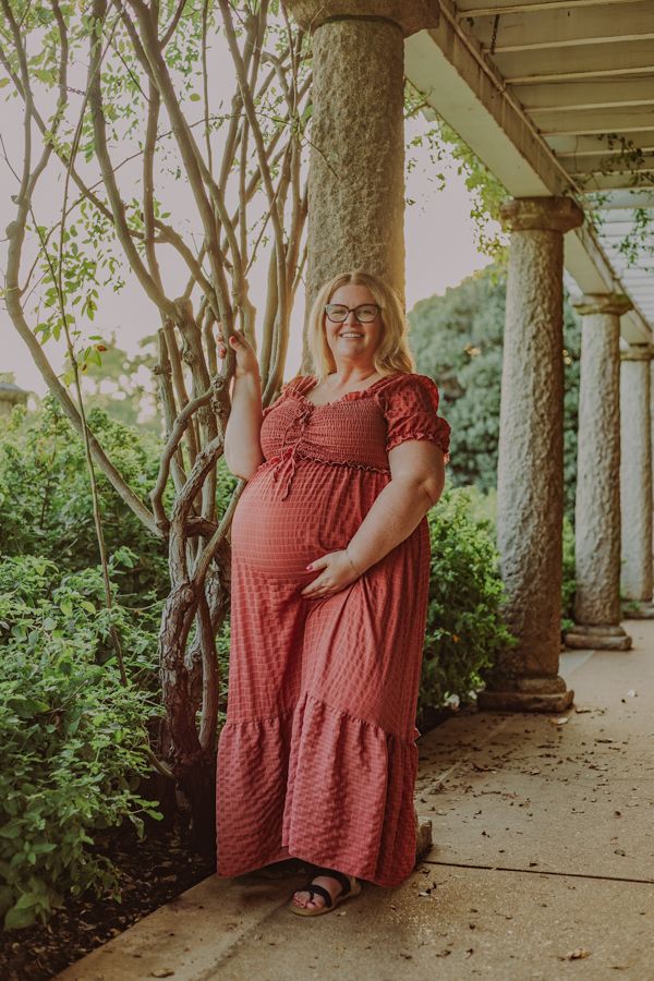 Smiling pregnant woman in pink maxi dress standing among columns and lush garden greenery.