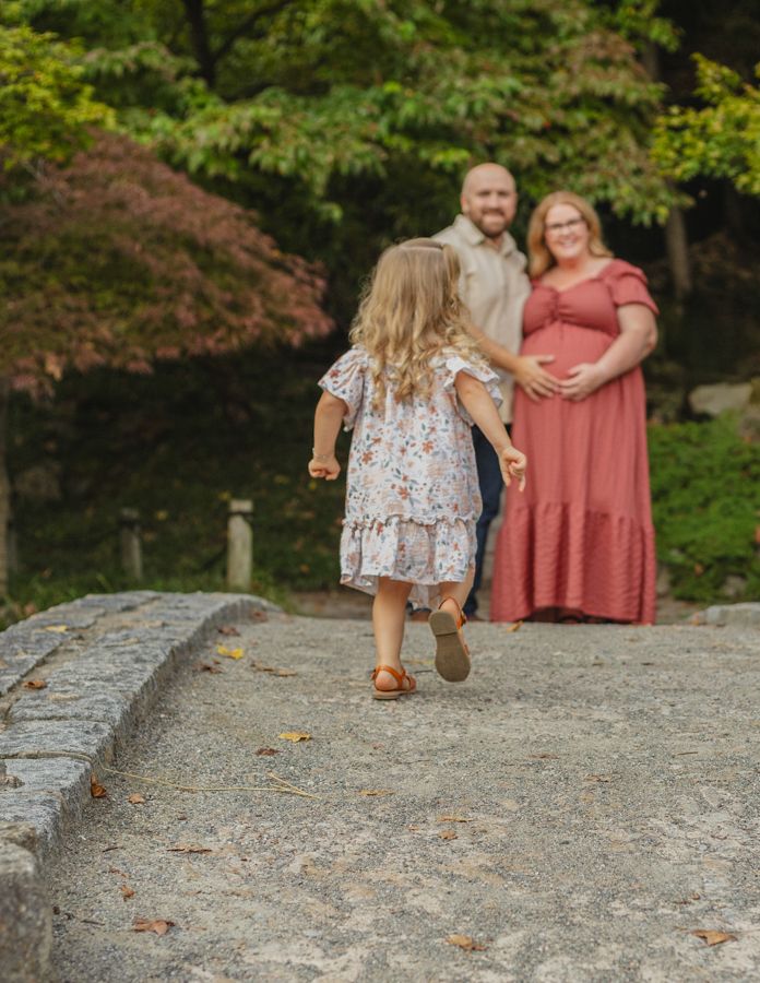 Young girl running toward her expecting parents on a stone bridge surrounded by lush green trees in a maternity shoot.