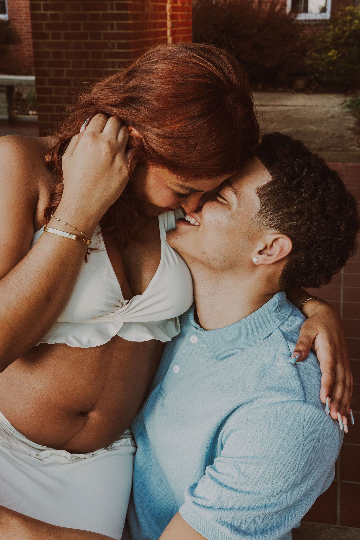 Pregnant couple sharing a tender kiss, woman in white crop top cradling baby bump outdoors.