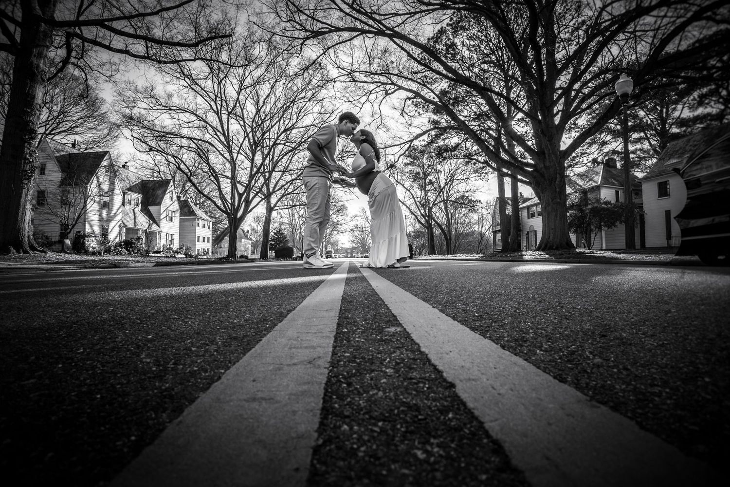 Black and white photo of couple sharing romantic dip kiss on tree-lined street in formal attire.