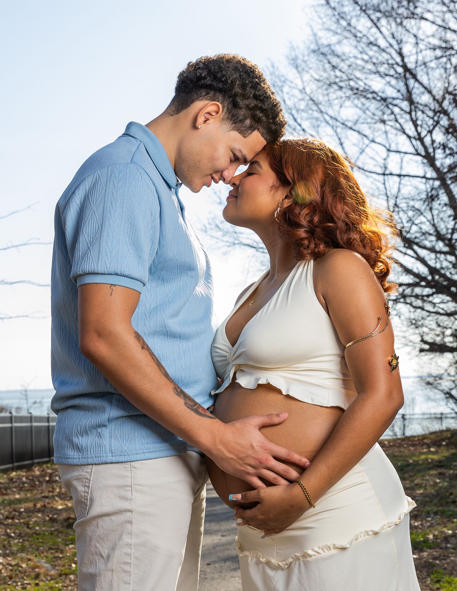 Expecting couple touching foreheads tenderly, hands on baby bump, in sunny outdoor park setting.