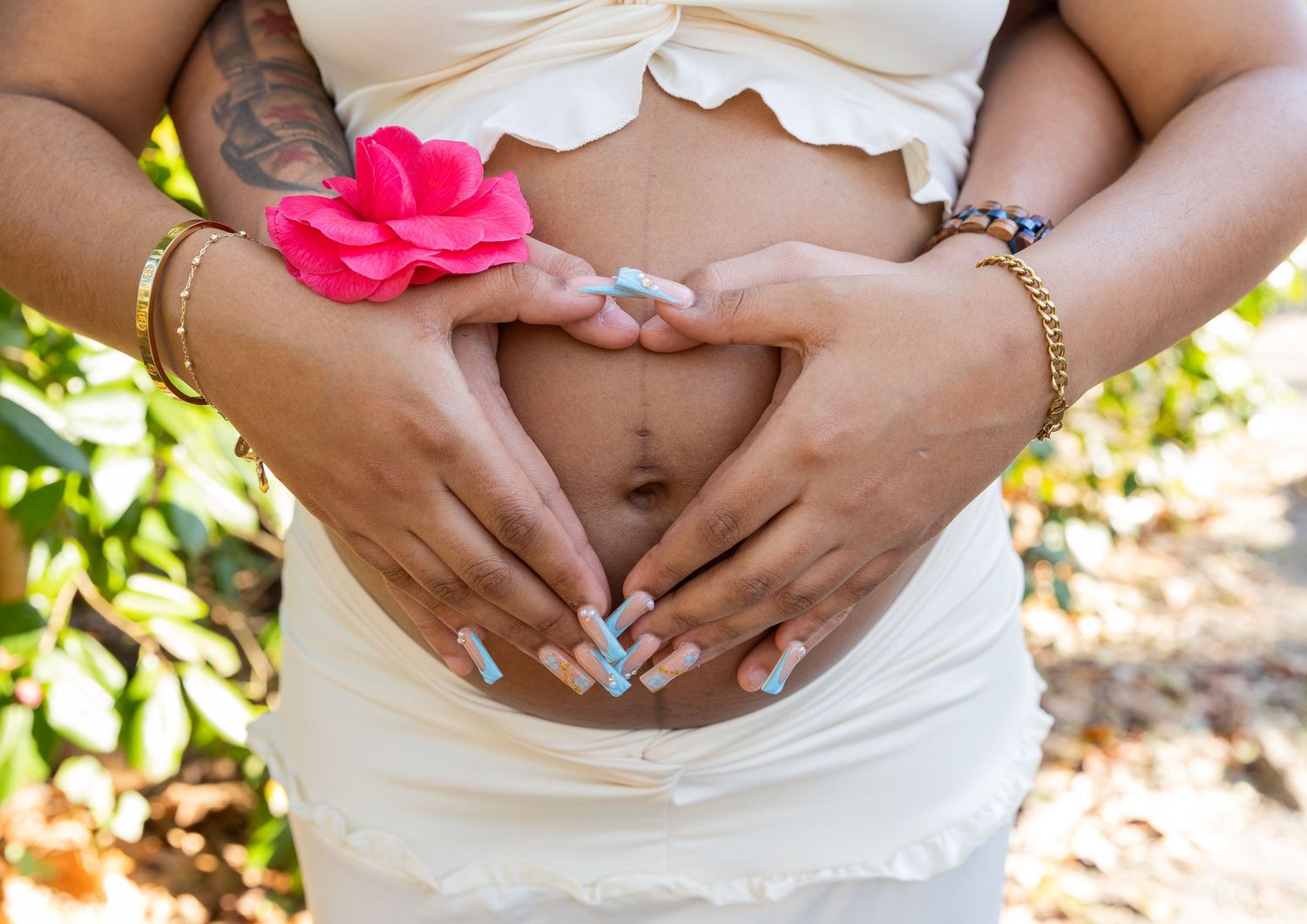 Close-up of pregnant belly with couple forming heart shape with hands, pink flower accent.