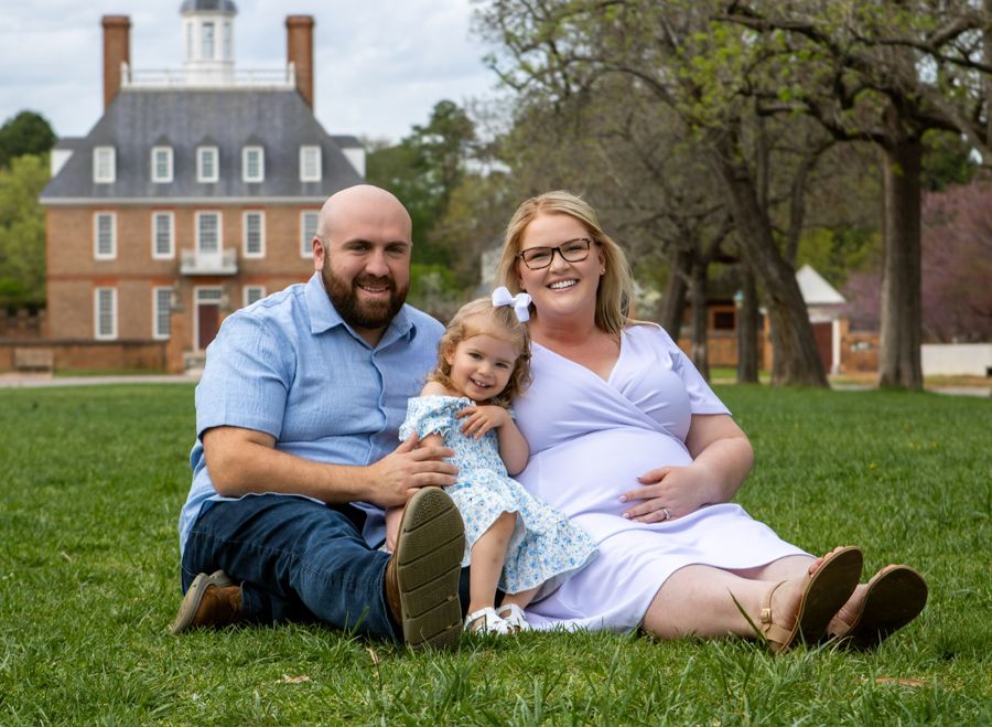 Happy family with toddler sitting on grass in front of a historic brick building during a spring maternity photo session.