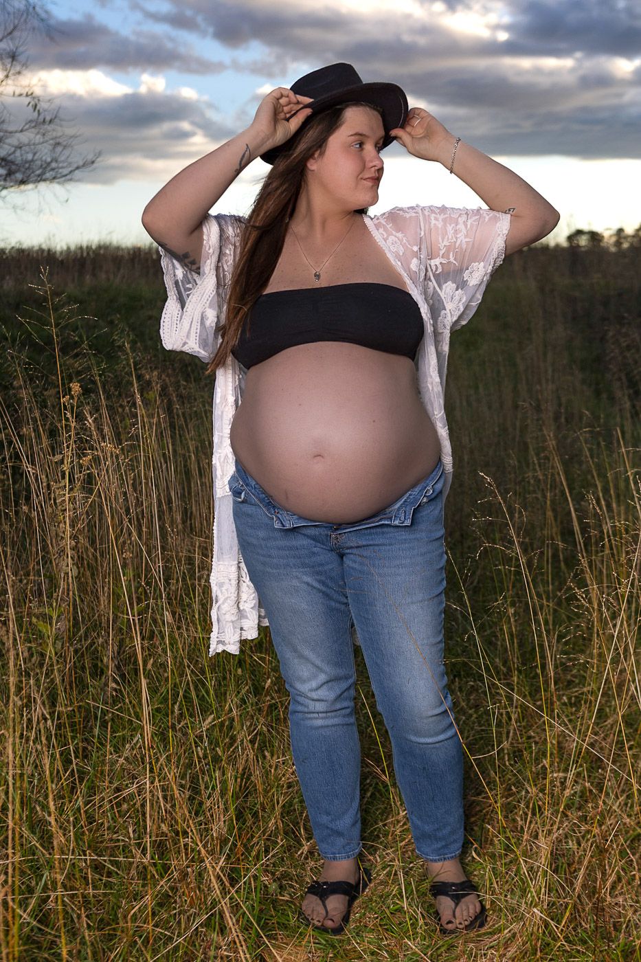 pregnant woman in a field holding hat wearing a black bra and white kimono with a dramatic sky behind