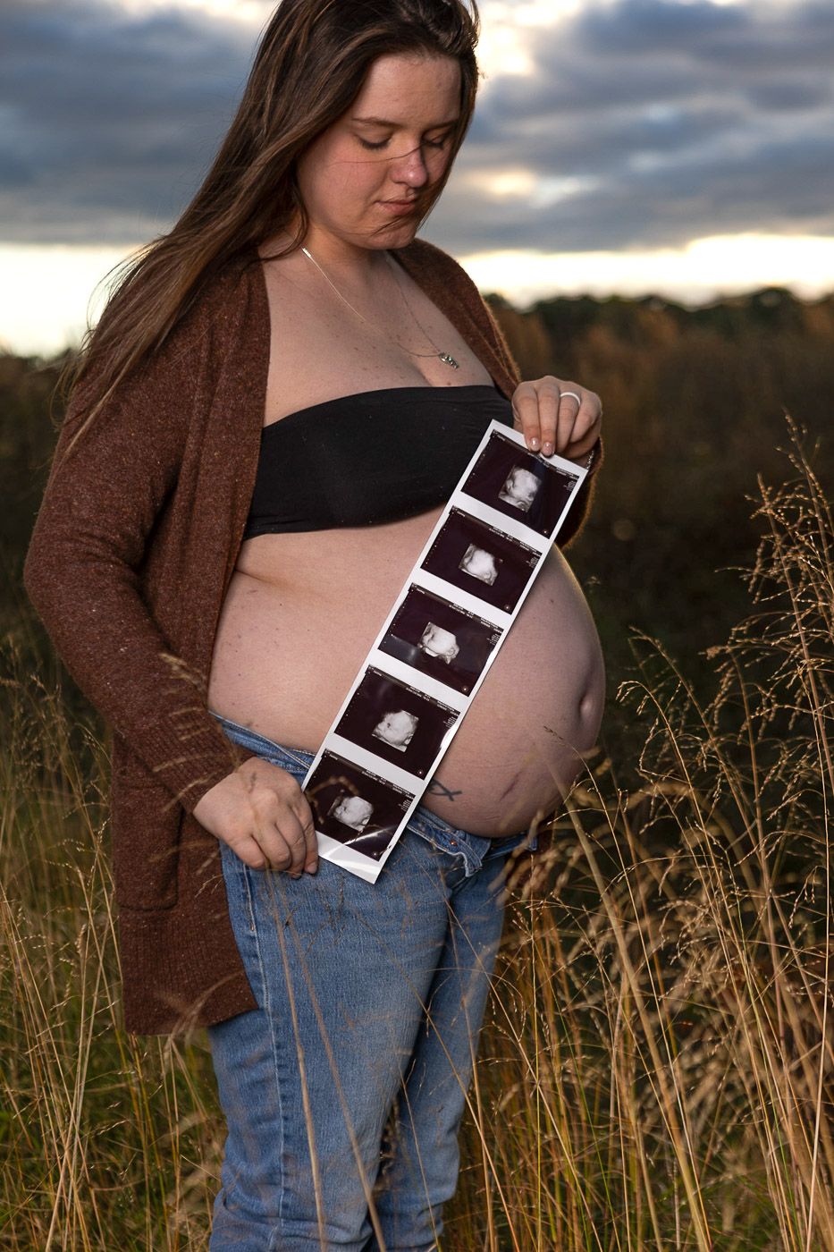 Pregnant woman in jeans and crop top holding ultrasound photos in a golden field at sunset, gazing at baby scans.