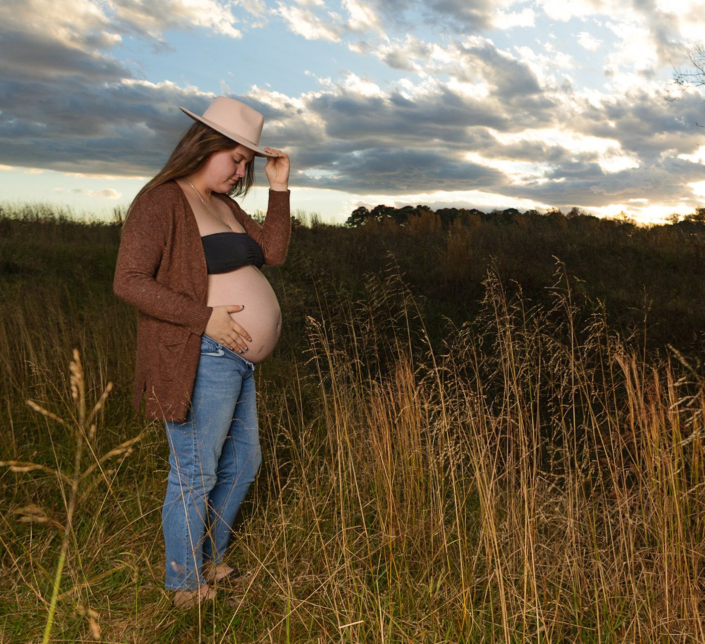 pregnant woman tipping her hat down, wearing a brown cardigan with a black bra and blue jeans, baby bump out in a field with a dramatic sky