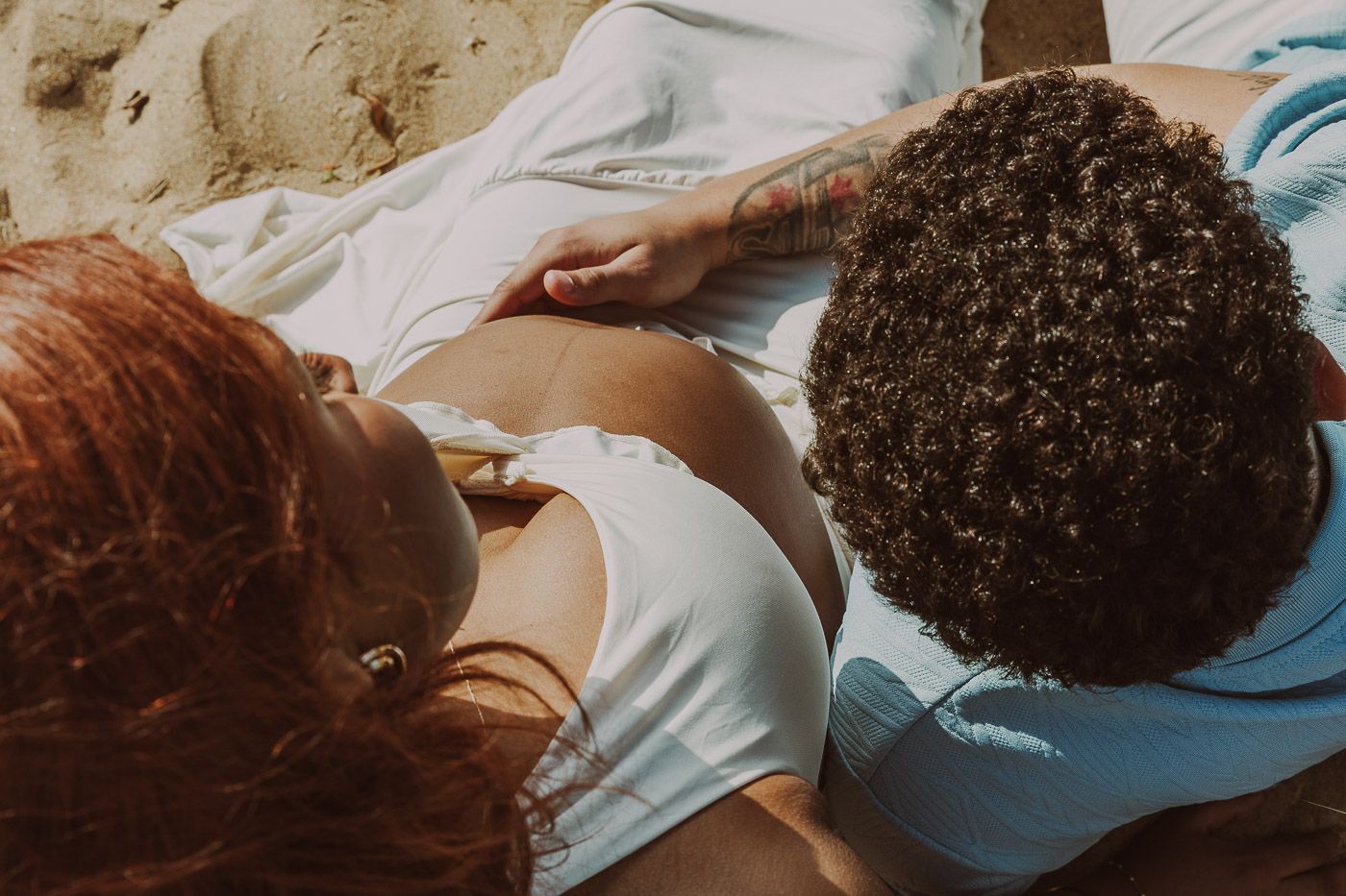 Overhead view of couple relaxing on beach sand, woman with red hair and tattooed partner in white shirts.