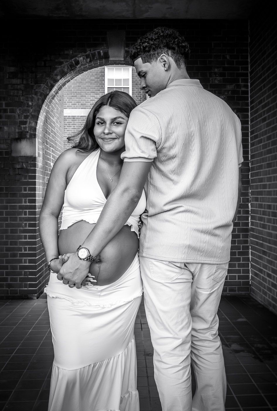 Black and white maternity photo of a couple in white outfits, man embracing pregnant woman against brick arch.