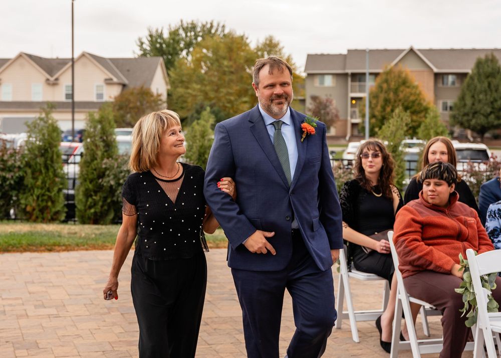 A man in a navy suit with an orange boutonniere walks arm-in-arm with his mom at an outdoor wedding ceremony at a Venue on the Ridge